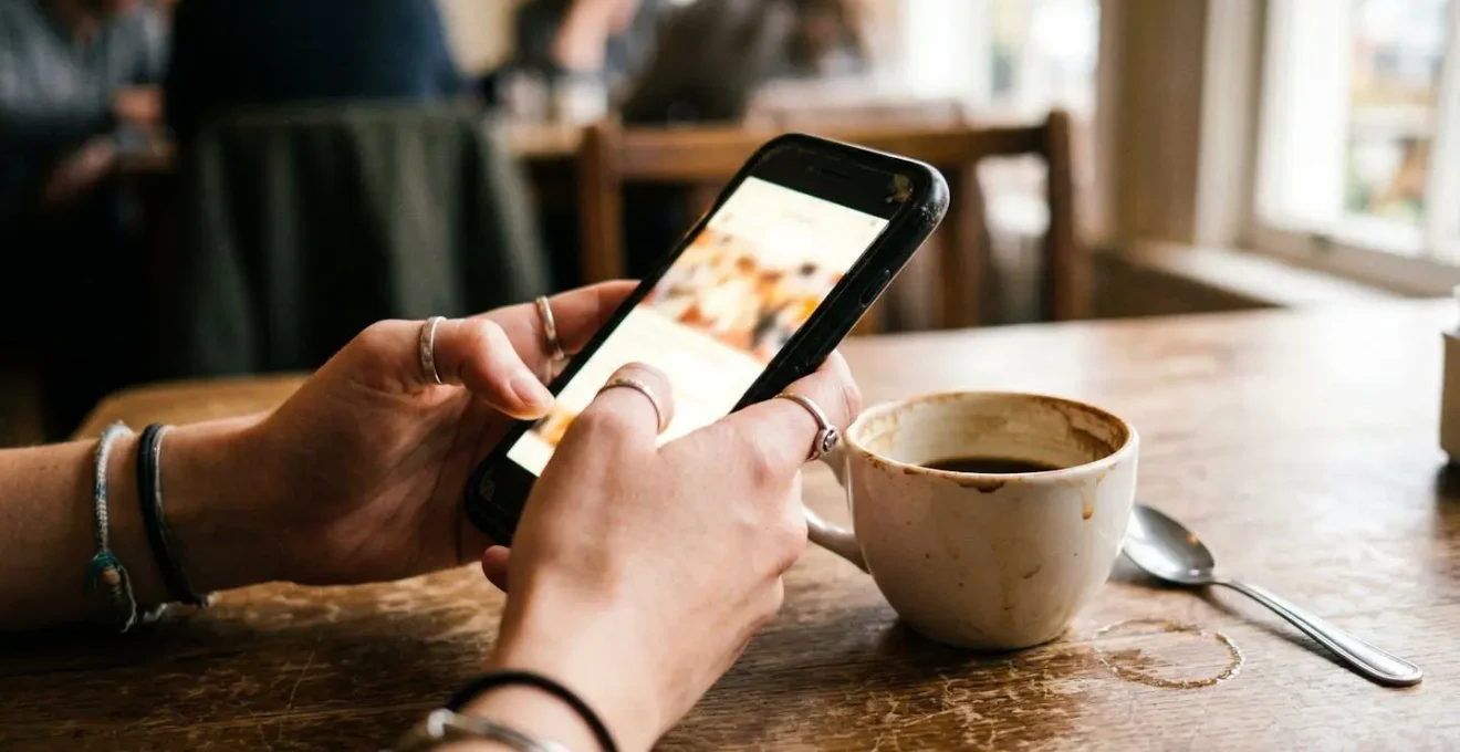 Mains tenant un smartphone face visible, café posé à côté sur une table en bois, éclairage naturel tamisé